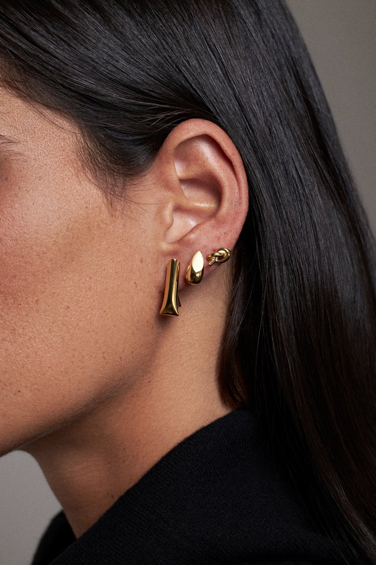 A close-up side of a CINCO girl with long dark hair wearing a pair of gold-toned ear cuff earrings from CINCO, including one flat vertical bar and one textured organic-shaped piece. Super cool for using everyday