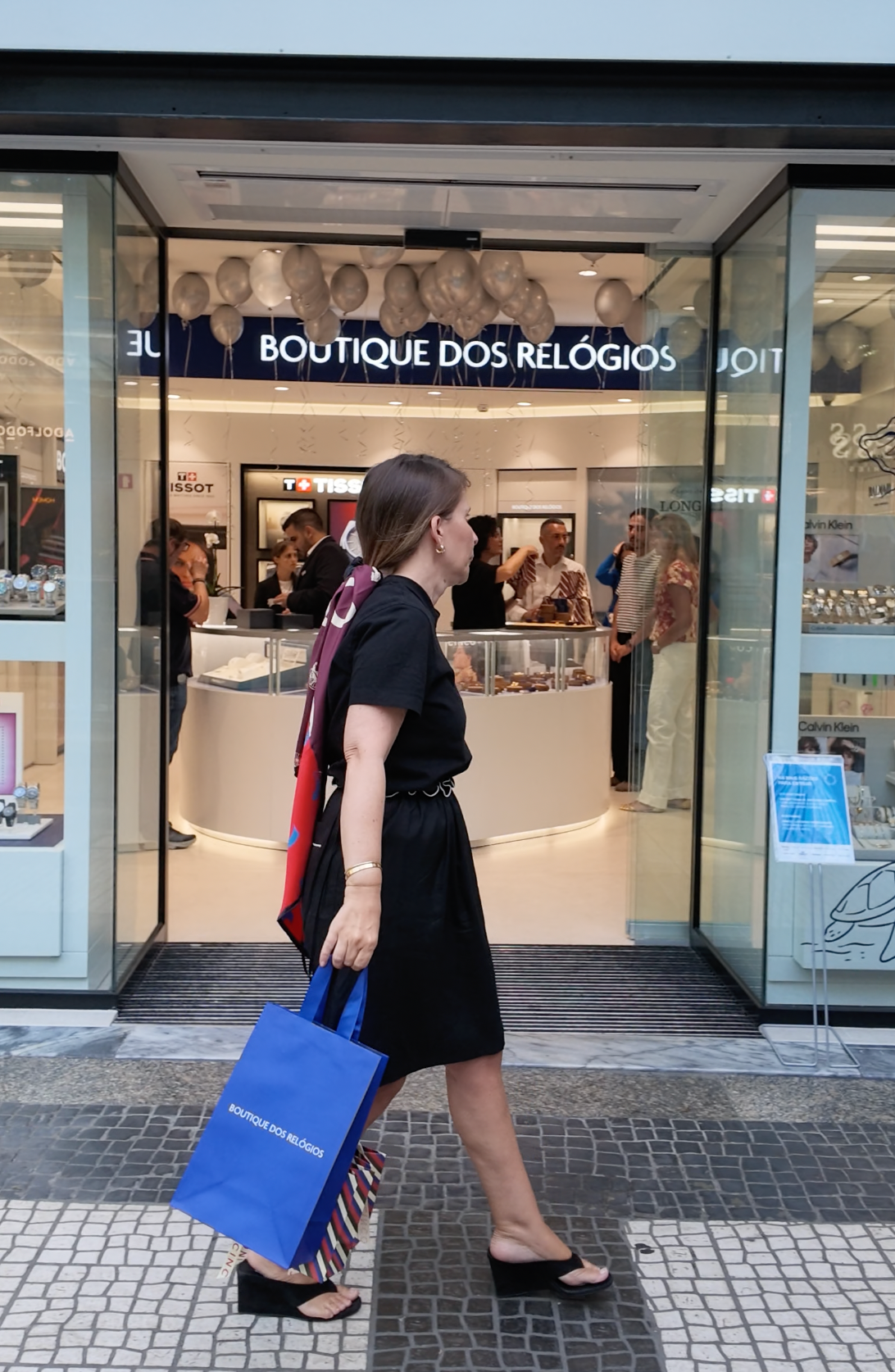 A woman in a black dress walks past a boutique storefront carrying a blue 'Boutique dos Relógios' shopping bag. The shop has large glass windows, interior display counters, and mannequins behind a sign reading 'BOUTIQUE DOS RELOGIOS'.