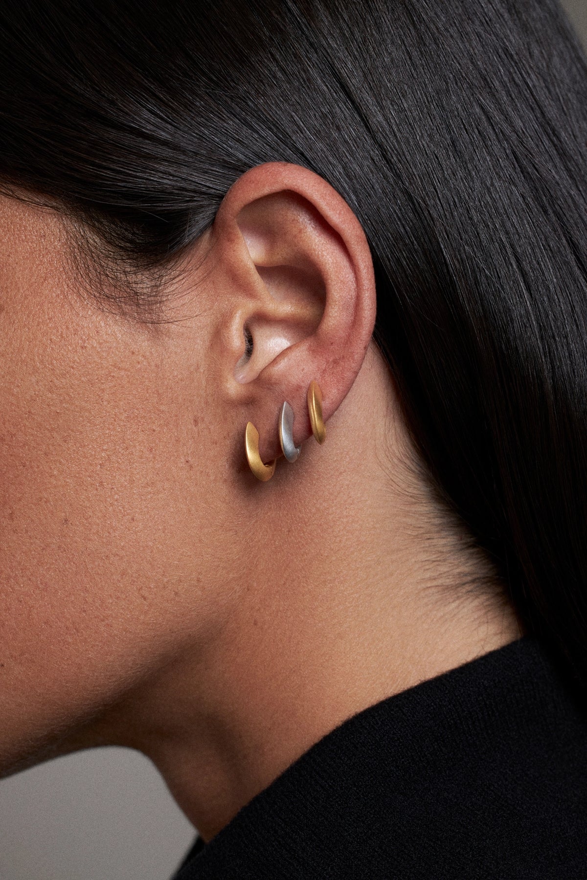 A close-up side profile of a CINCO girl wearing three small crescent-shaped hoop earrings in gold, silver, and rose gold on her ear. Super cool look for a night out.