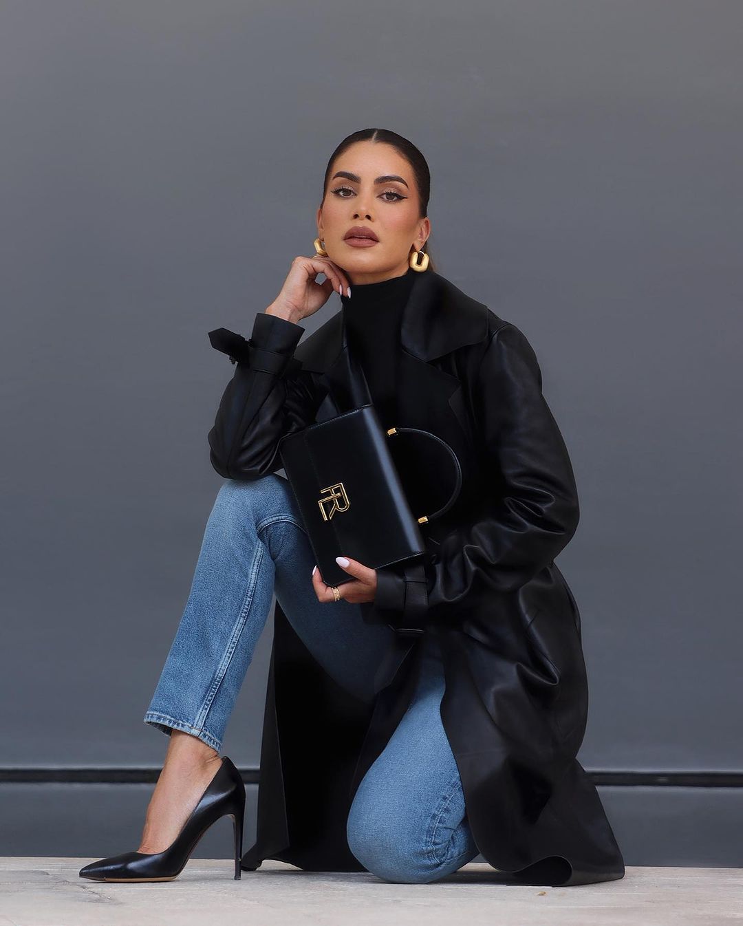 Woman in black leather coat crouching with a black handbag, showcasing elegant style and black high heels.