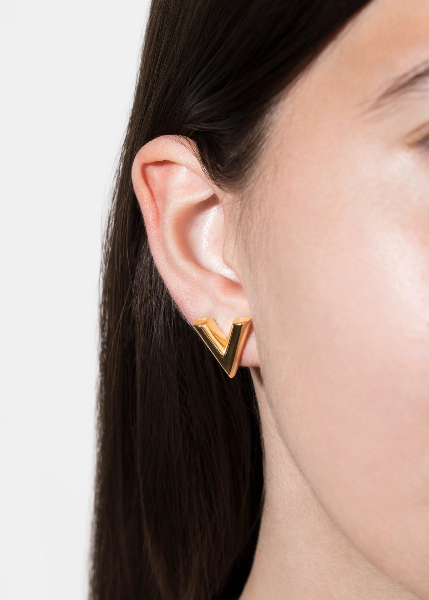 A close-up of a model's ear wearing a gold geometric V-shaped stud earring against a light gray background.